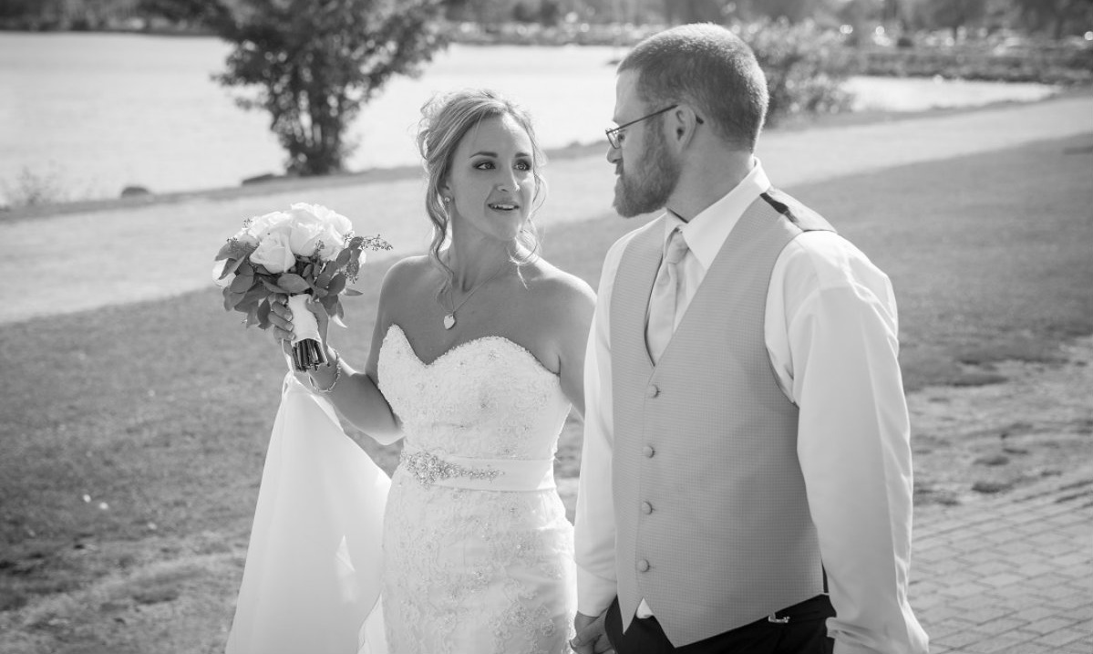 A large group of wedding participants pose outdoors while the couple kisses in the middle